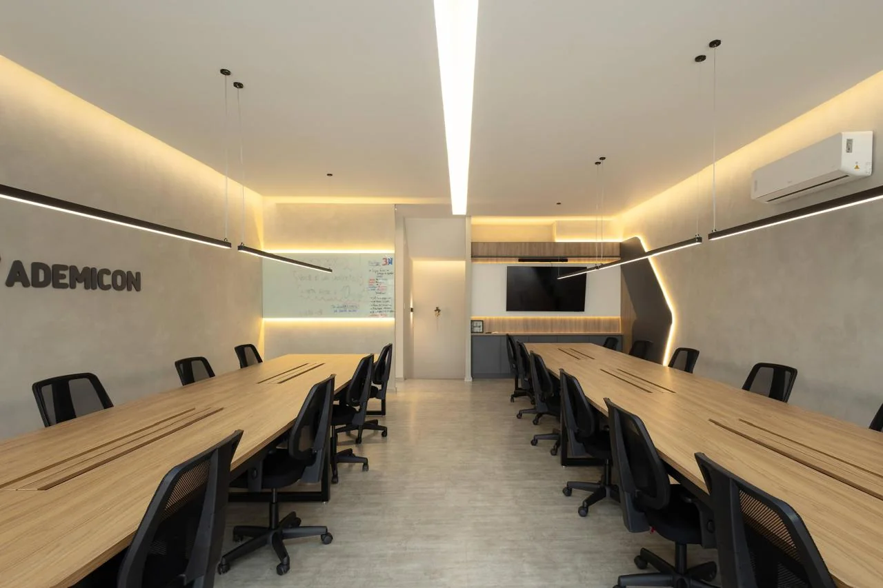 Sala ampla com mesas corporativas, iluminação linear e logo Ademicon na parede